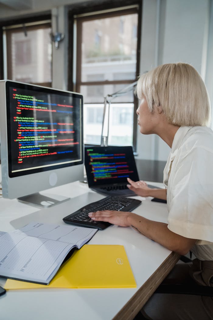 Woman with blonde hair programming on a computer in a modern office, focused on multicolored script on screen.