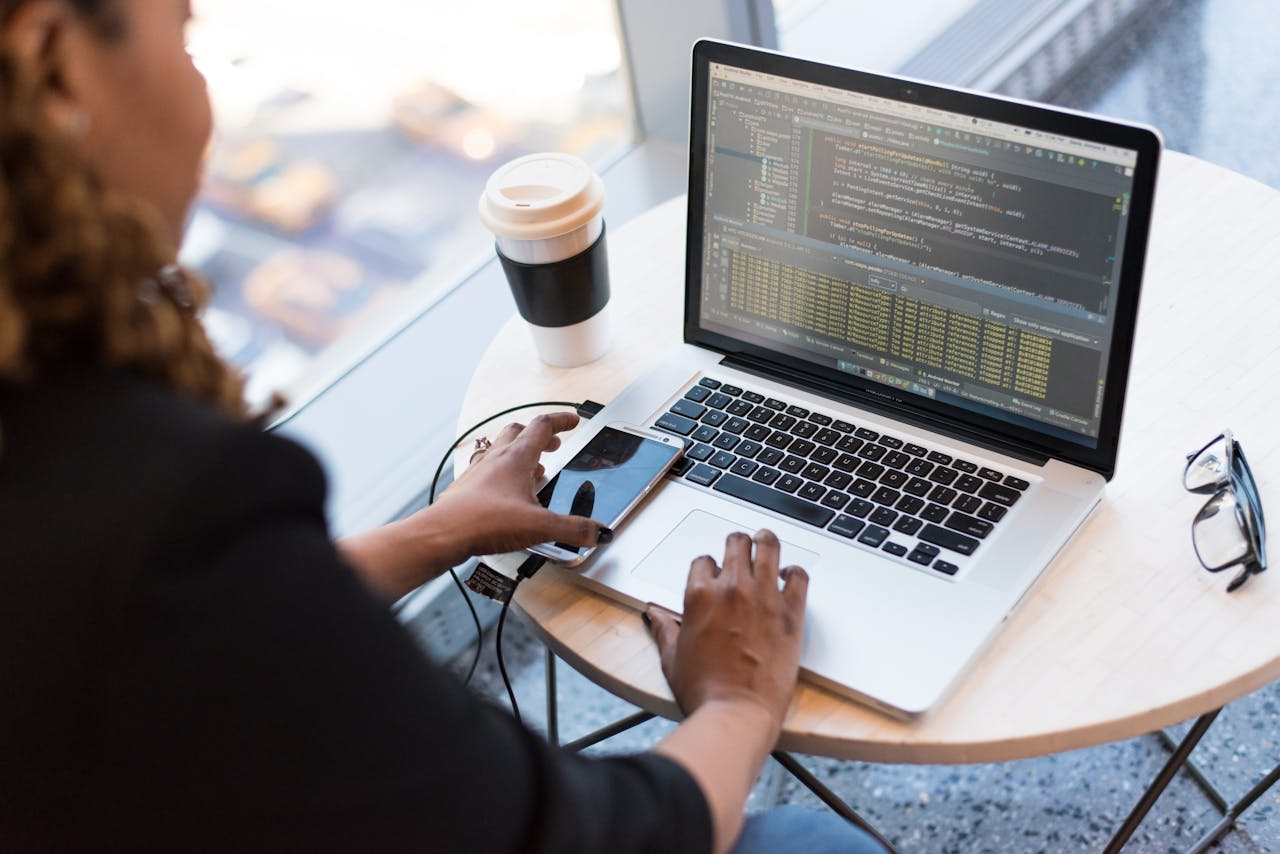 who-we-are Black woman programming on a laptop with coffee, smartphone, and glasses on a desk in an office.